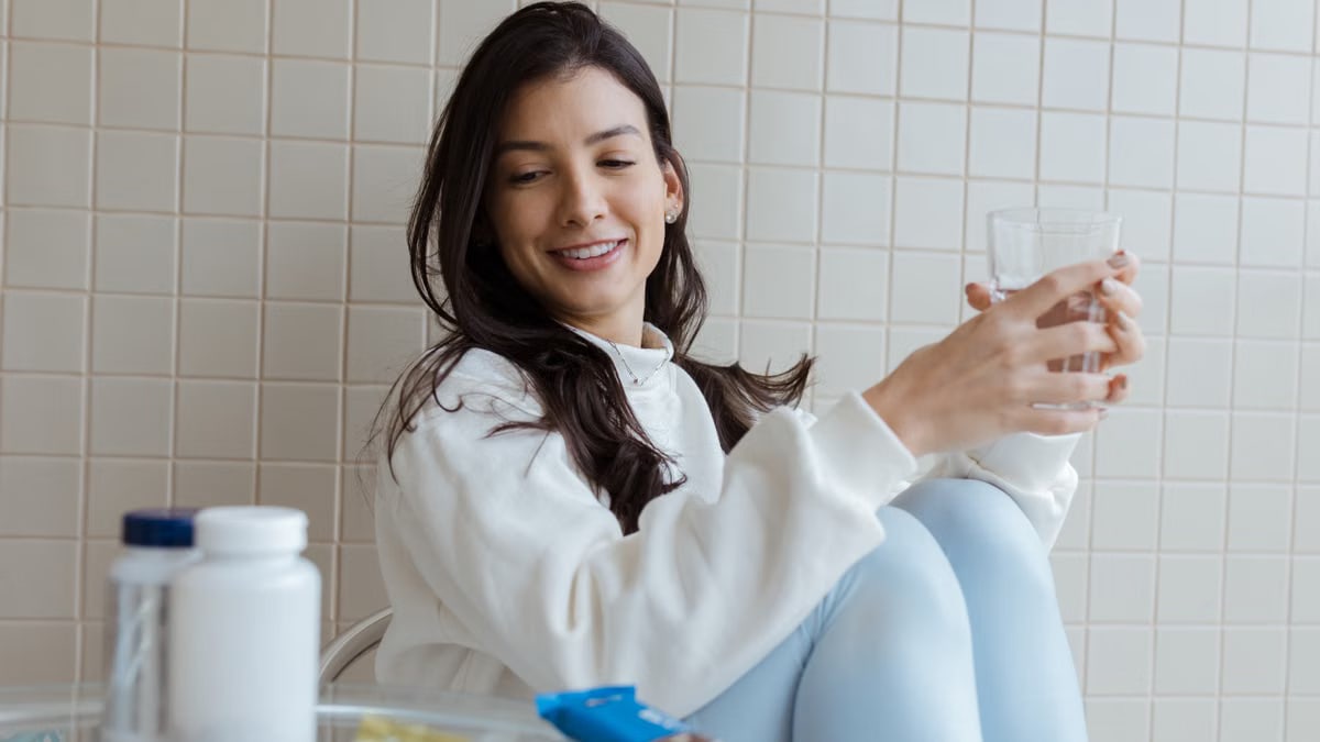 Smiling woman holding a glass while seated, with pill bottles on the table.